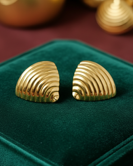 Gold shell-shaped earrings on a green velvet cushion with gold objects in the background.