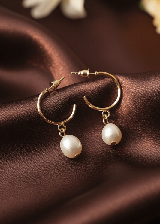 Gold hoop earrings with pearl drops on a brown satin surface with white flowers in the background.