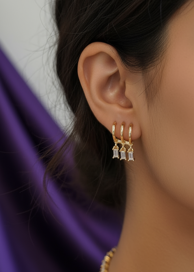 Close-up of a person wearing gold hoop earrings with small stones against a blurred purple fabric background.